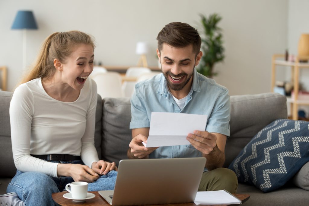 Photo of happy, ecstatic couple looking at document while sitting on couch behind a coffee table with a laptop computer on it.