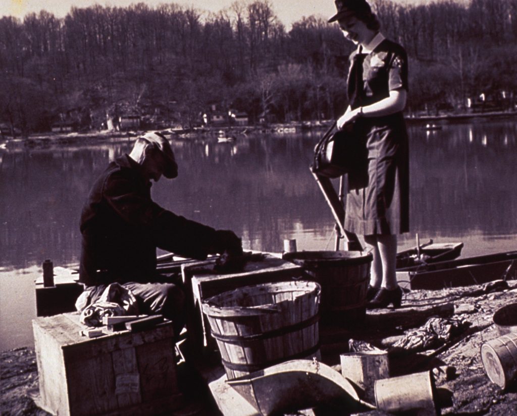 Public health nurse standing while visiting patient sitting on river shore sorting fish in barrels.